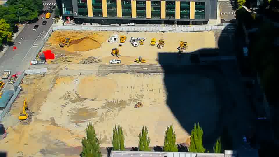 Construction camera frame at 12:15 PM PT. A small white vehicle is now visible in the central area of the site, having appeared between these frames.