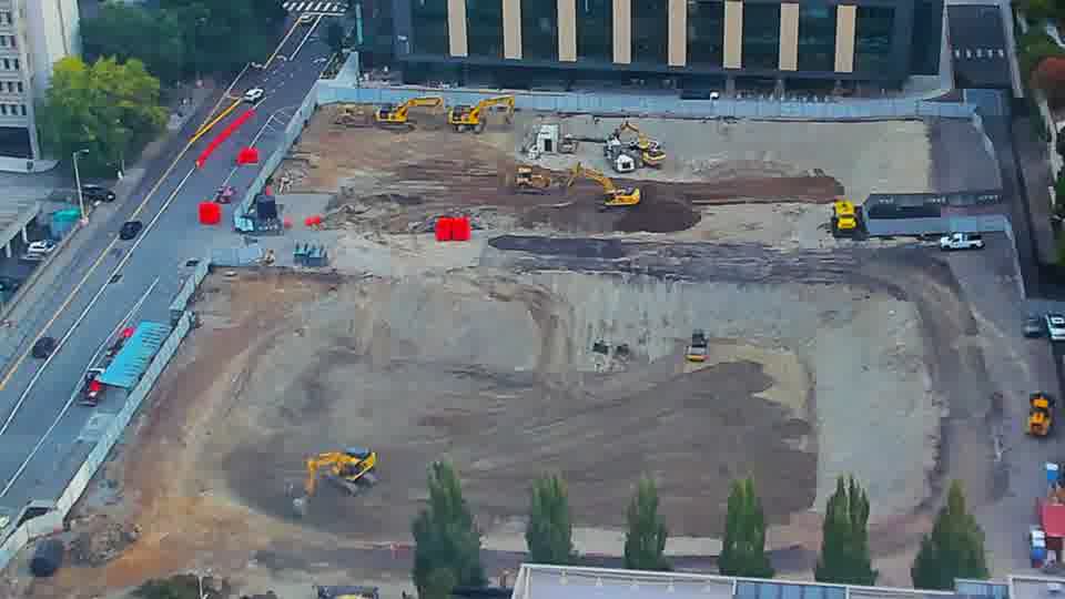 Construction camera frame at 7:45 AM PT. The red artifact obscuring the view in the earlier frame is gone, revealing activity across the site. A small orange vehicle has moved from the lower-right to the center of the block.