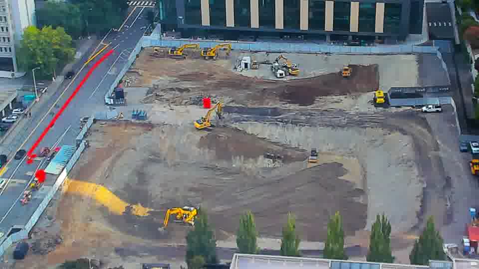 Construction camera frame at 8:30 AM PT. A yellow machine has moved from the lower-left to the center of the block. A truck has entered the site from the driveway on the left.