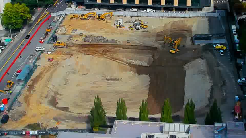 Construction camera frame at 12:00 PM PT. A small white vehicle has entered the site from the driveway on the left side of the frame. Otherwise, the scene appears unchanged.