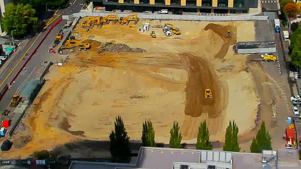 Construction camera frame at 1:15 PM PT. A piece of heavy machinery has moved slightly within the central area of the site. Otherwise, the scene appears unchanged.