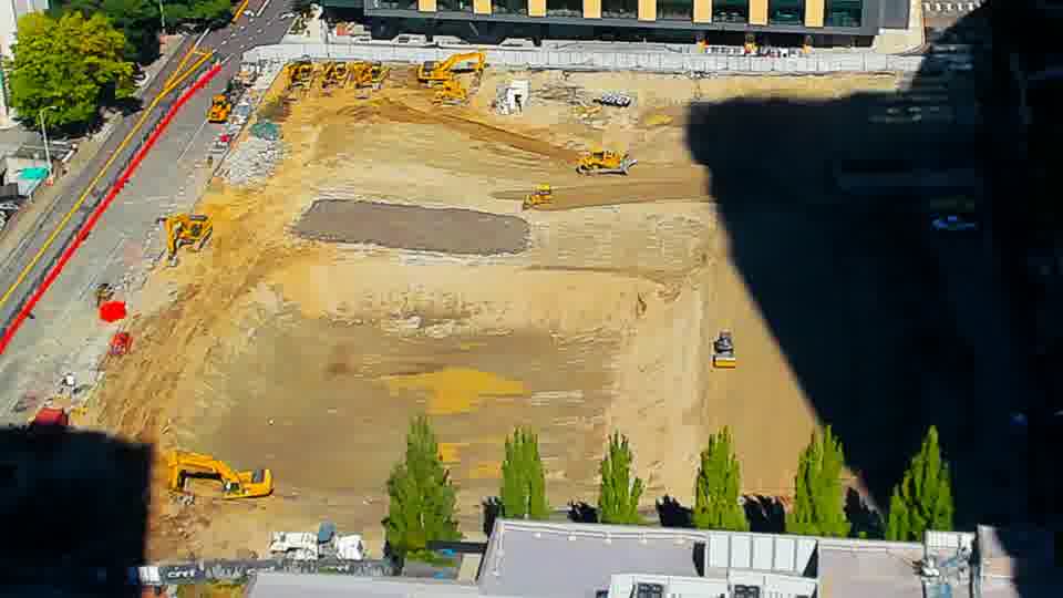 Construction camera frame at 12:15 PM PT. A small yellow vehicle has moved from the lower-right quadrant to the center of the site.